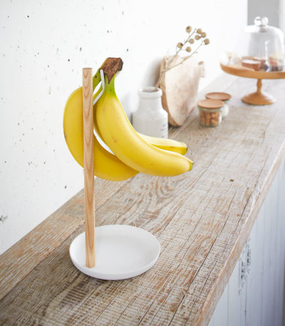 Angled view of the Yamazaki Home Banana Stand - Steel + Wood on a rustic wooden kitchen counter, with yellow bananas suspended above its white dish-shaped base.