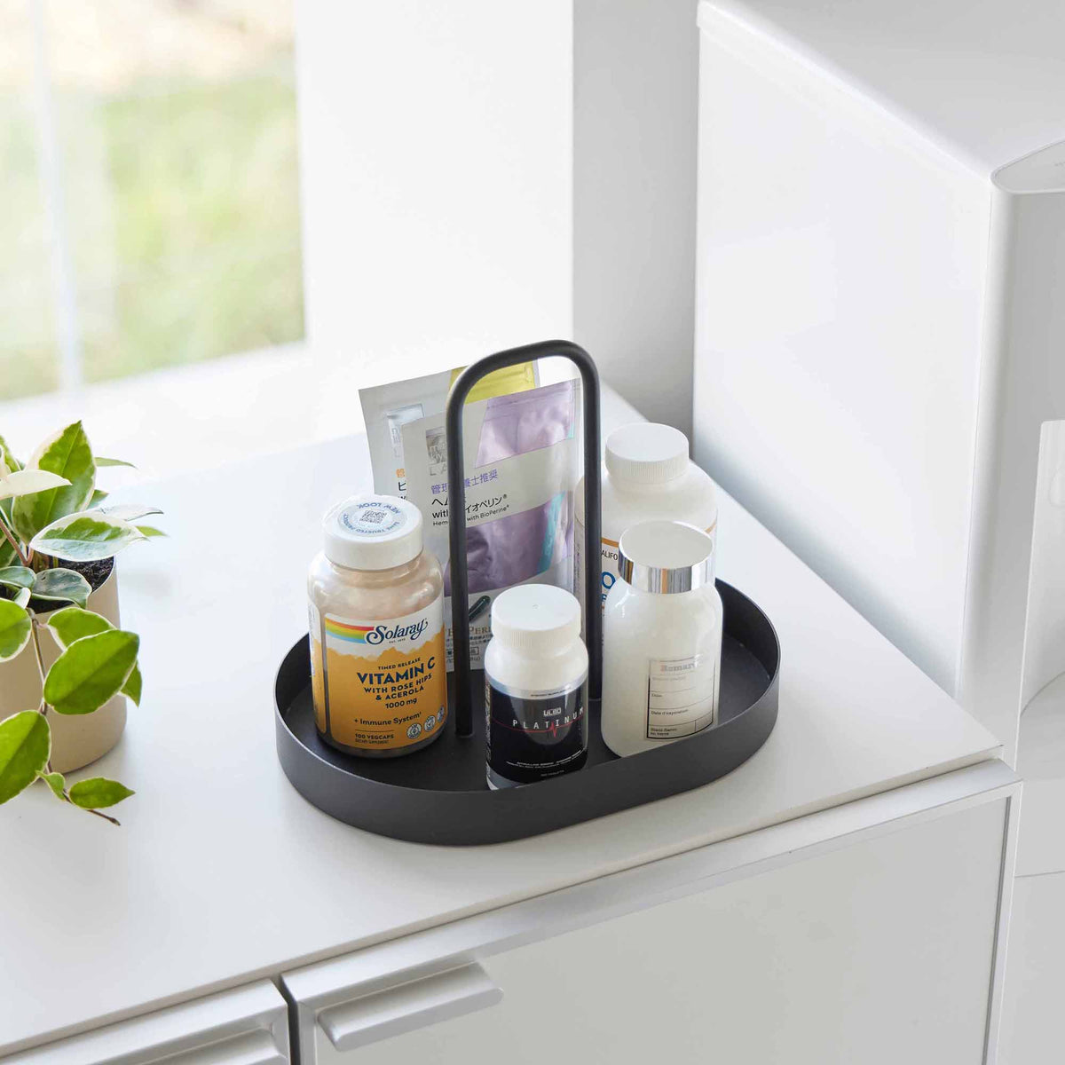 Angled view of the black Yamazaki Home Vanity Tray with Handle - Steel filled with bottles and packets on a white cabinet near a bright window.