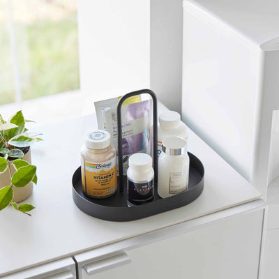 Angled view of the black Yamazaki Home Vanity Tray with Handle - Steel filled with bottles and packets on a white cabinet near a bright window.