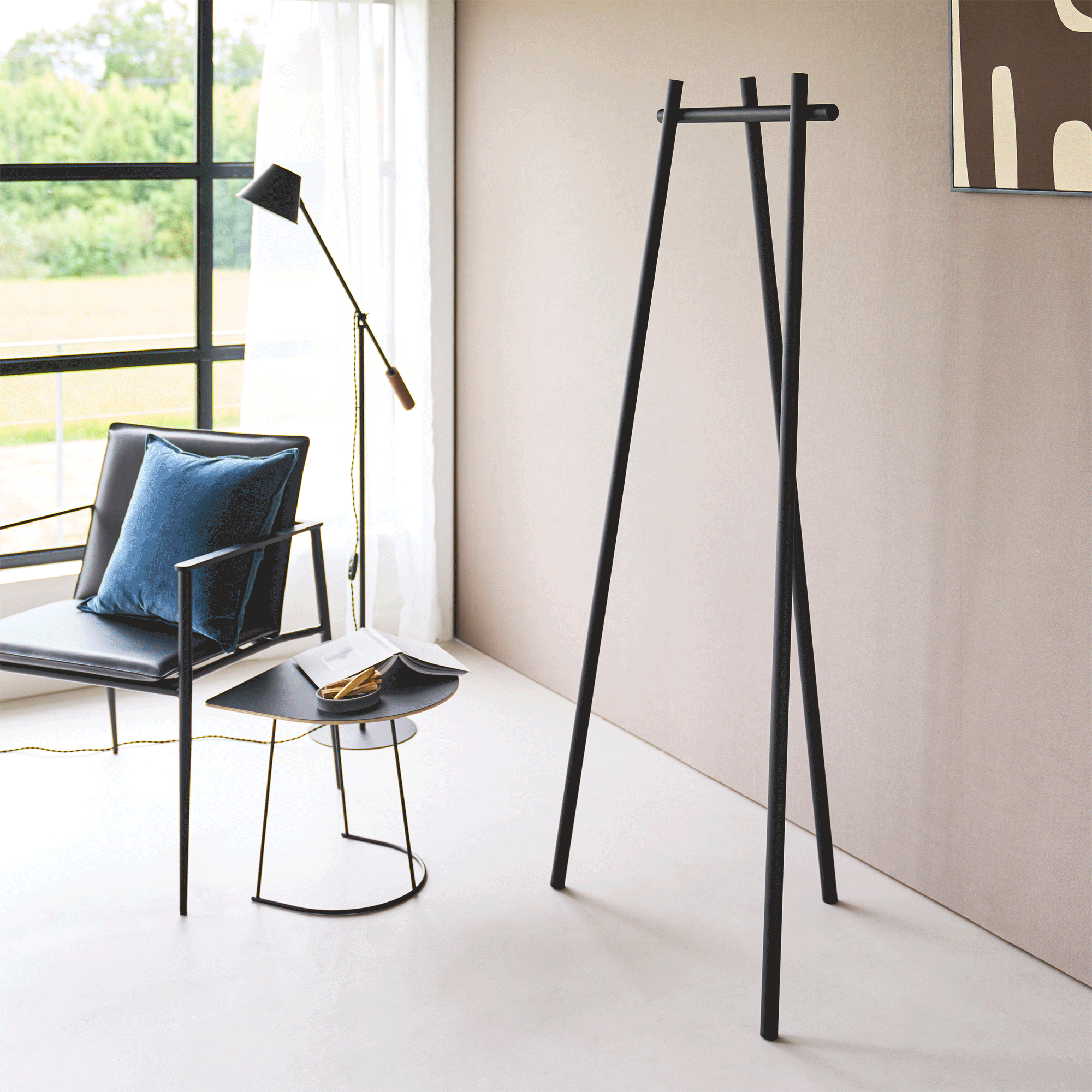 Black Yamazaki Home Tri-Post Coat Rack (60" H) - Steel holding dark and olive-brown outerwear, a neutral hat and mustard shoulder bag beside a window and black chair with a blue cushion.