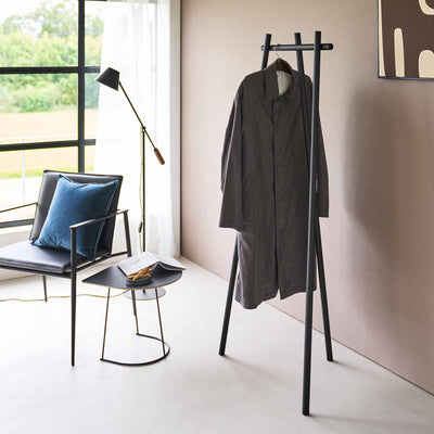 Black Yamazaki Home Tri-Post Coat Rack (60" H) - Steel holding a single long charcoal coat on a wooden hanger against a beige wall, with its splayed legs visible below.