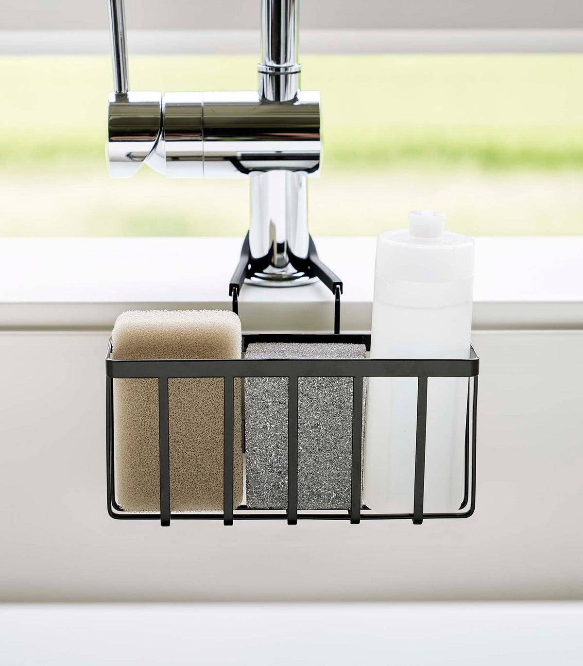 Front view of the black Yamazaki Home Faucet-Hanging Sponge Caddy - Steel beneath a chrome faucet, with beige and grey sponges standing beside a white bottle.