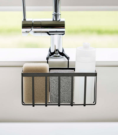 Front view of the black Yamazaki Home Faucet-Hanging Sponge Caddy - Steel beneath a chrome faucet, with beige and grey sponges standing beside a white bottle.