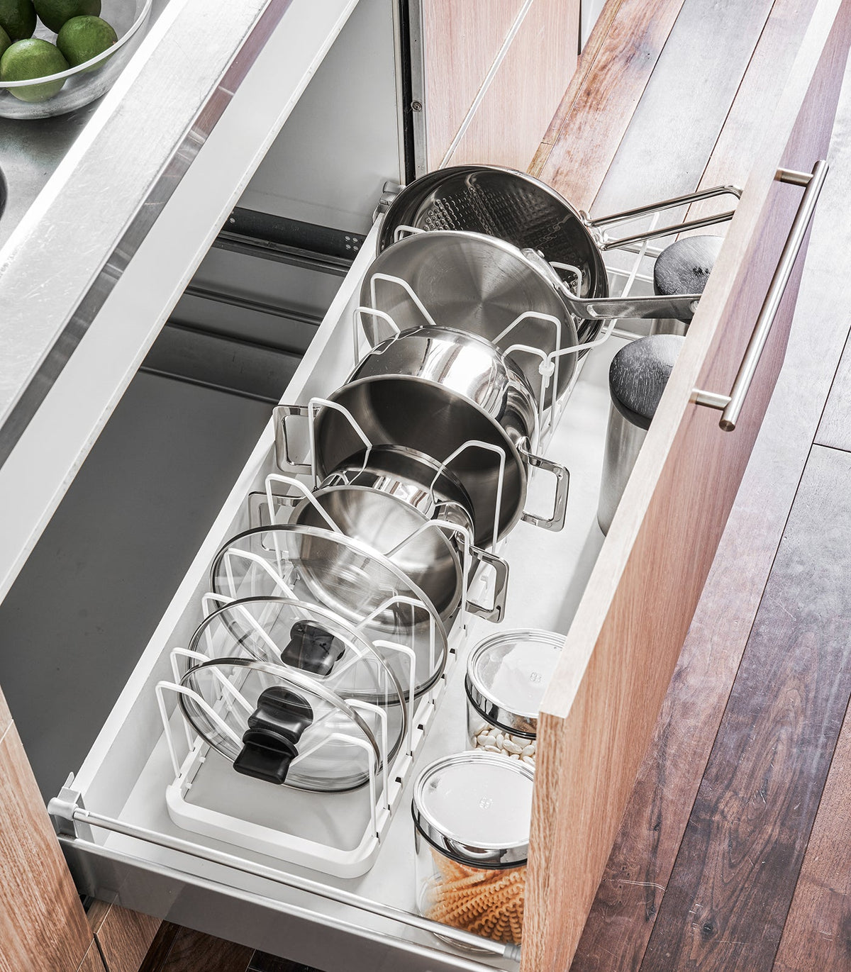 White Yamazaki Home Adjustable Pot Lid Organizer - Steel inside an open kitchen drawer, separating pans and lids beside cylindrical storage containers.