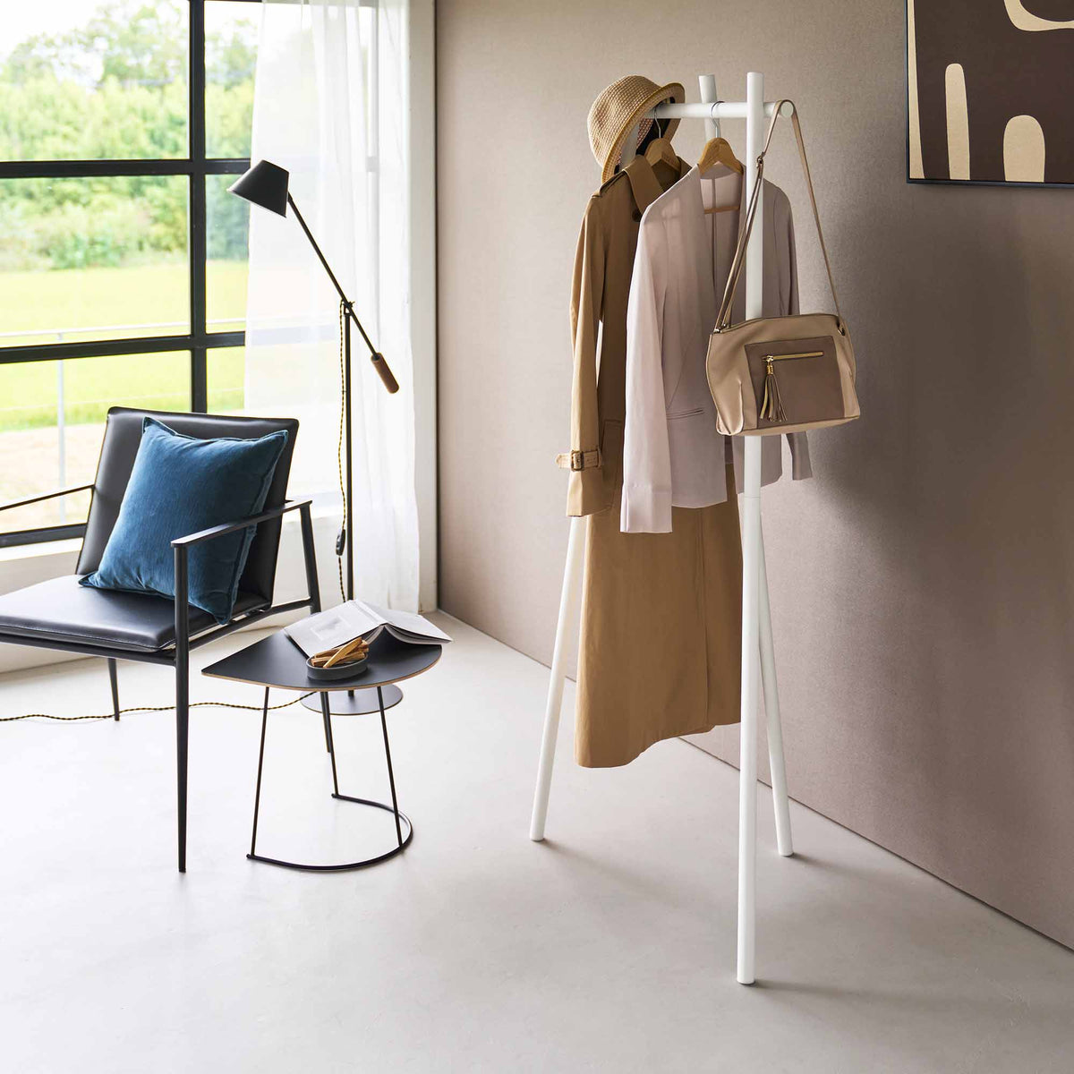 White Yamazaki Home Tri-Post Coat Rack (60" H) - Steel holding layered tan and pale clothing, a straw hat and a small beige shoulder bag in a bright sitting area.