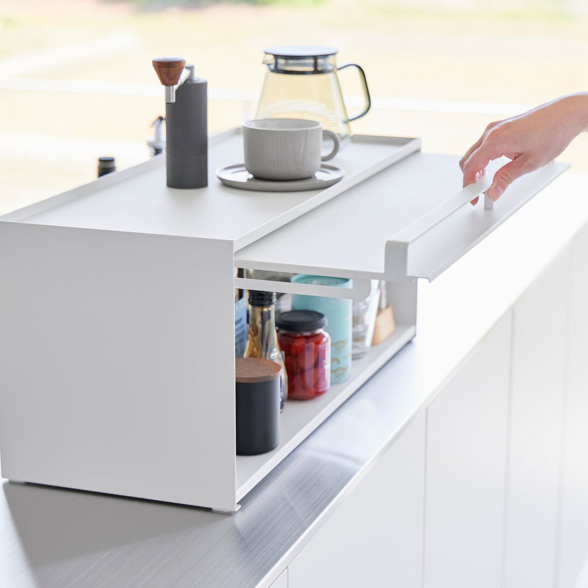 A hand raises the white front panel of the Yamazaki Home Open-Back Countertop Storage Rack - Steel, exposing bottles, jars and containers stored inside.