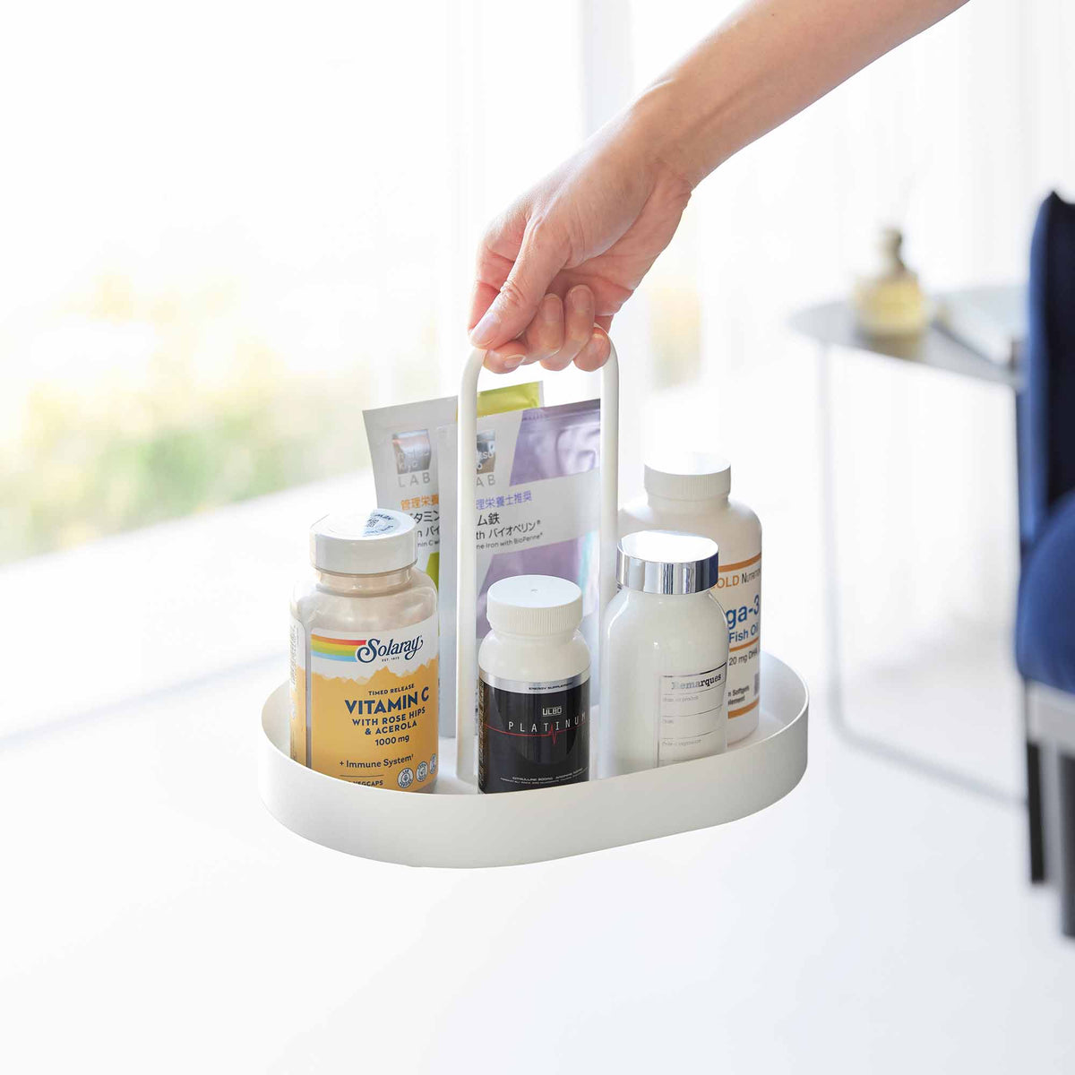 A person carries the white Yamazaki Home Vanity Tray with Handle - Steel by its central handle, with bottles and packets standing inside the raised rim.