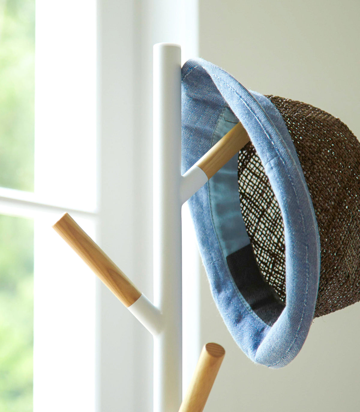 Upper section of the white Yamazaki Home Coat Rack (70" H) - Steel beside a window, with a blue-trimmed woven hat hanging from one wood-tipped branch.
