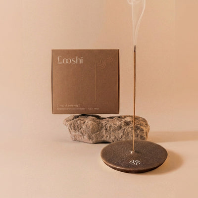 Burning incense stick releasing a thin trail of smoke from a dark brown Looshi Ring of Serenity ceramic holder, displayed with natural stone and packaging