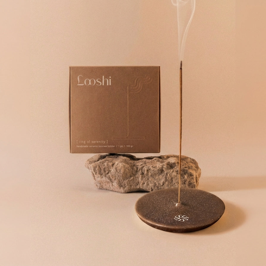 Burning incense stick releasing a thin trail of smoke from a dark brown Looshi Ring of Serenity ceramic holder, displayed with natural stone and packaging