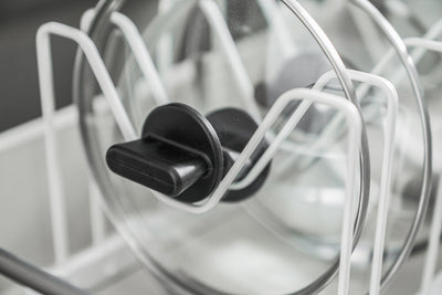Detail of a glass lid in the white Yamazaki Home Adjustable Pot Lid Organizer - Steel, showing its black handle resting within a dip in the wire divider.