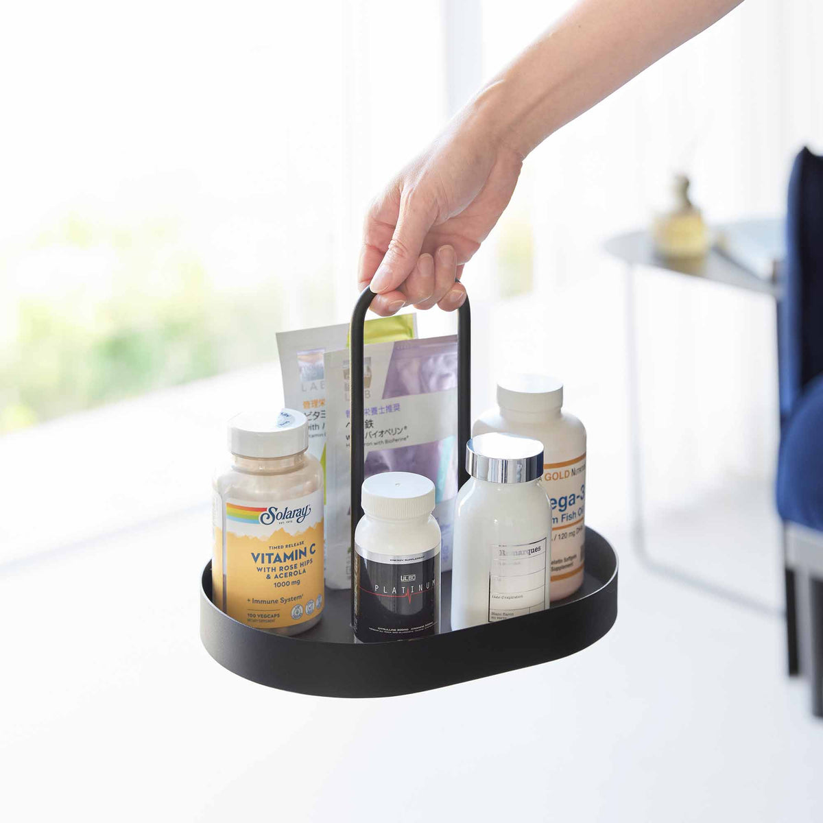 A hand lifts the black Yamazaki Home Vanity Tray with Handle - Steel by its rounded loop handle, carrying bottles and packets together.