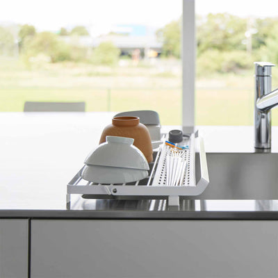 End view of the white Yamazaki Home Slim Over-the-Sink Dish Drying Rack - Steel spanning a sink, with bowls and cups arranged beside the perforated utensil section near a bright window.