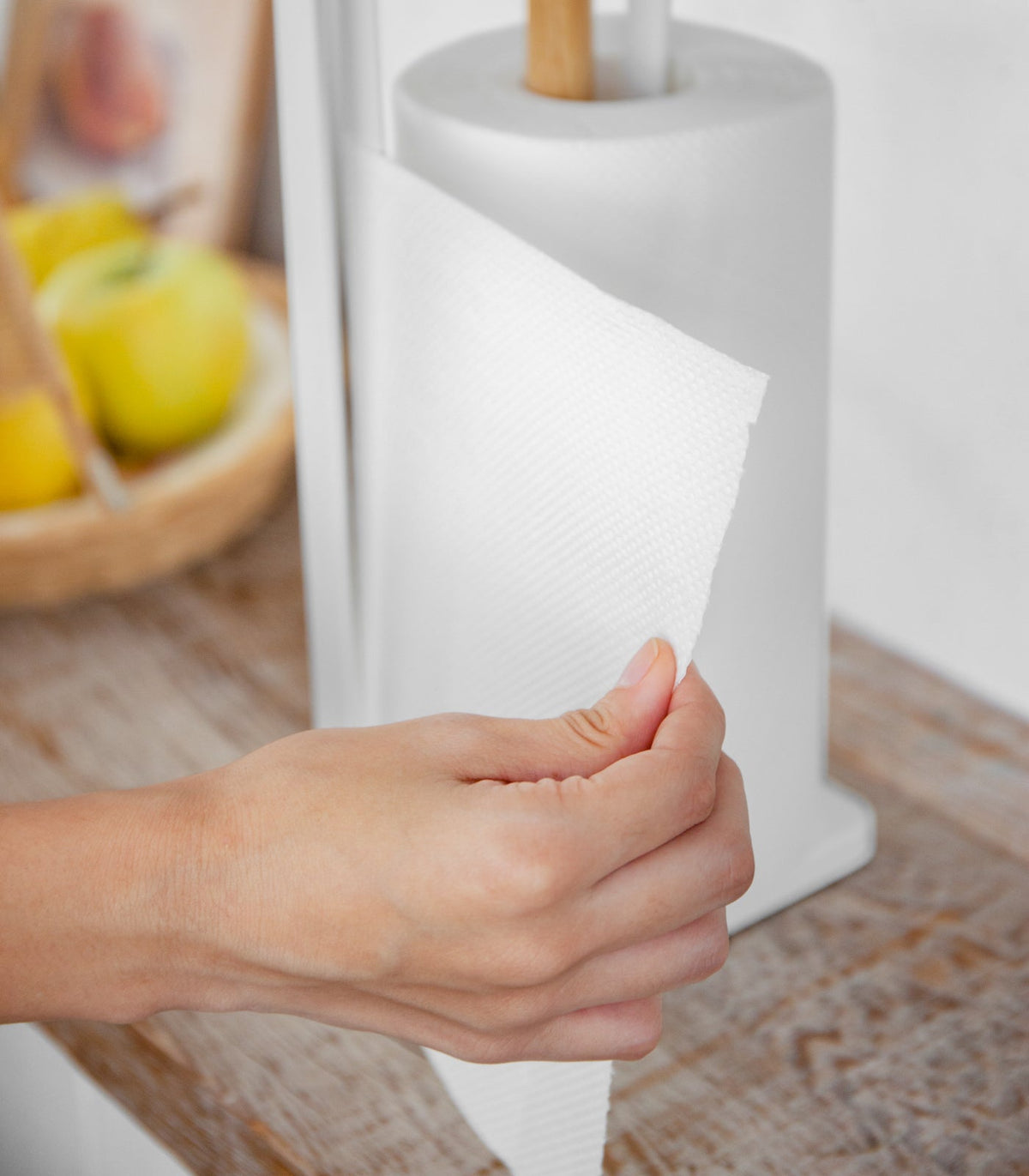A hand grips the loose paper towel beside the Yamazaki Home Paper Towel Holder - Steel + Wood, showing the sheet drawn across the slim white upright on a kitchen counter.