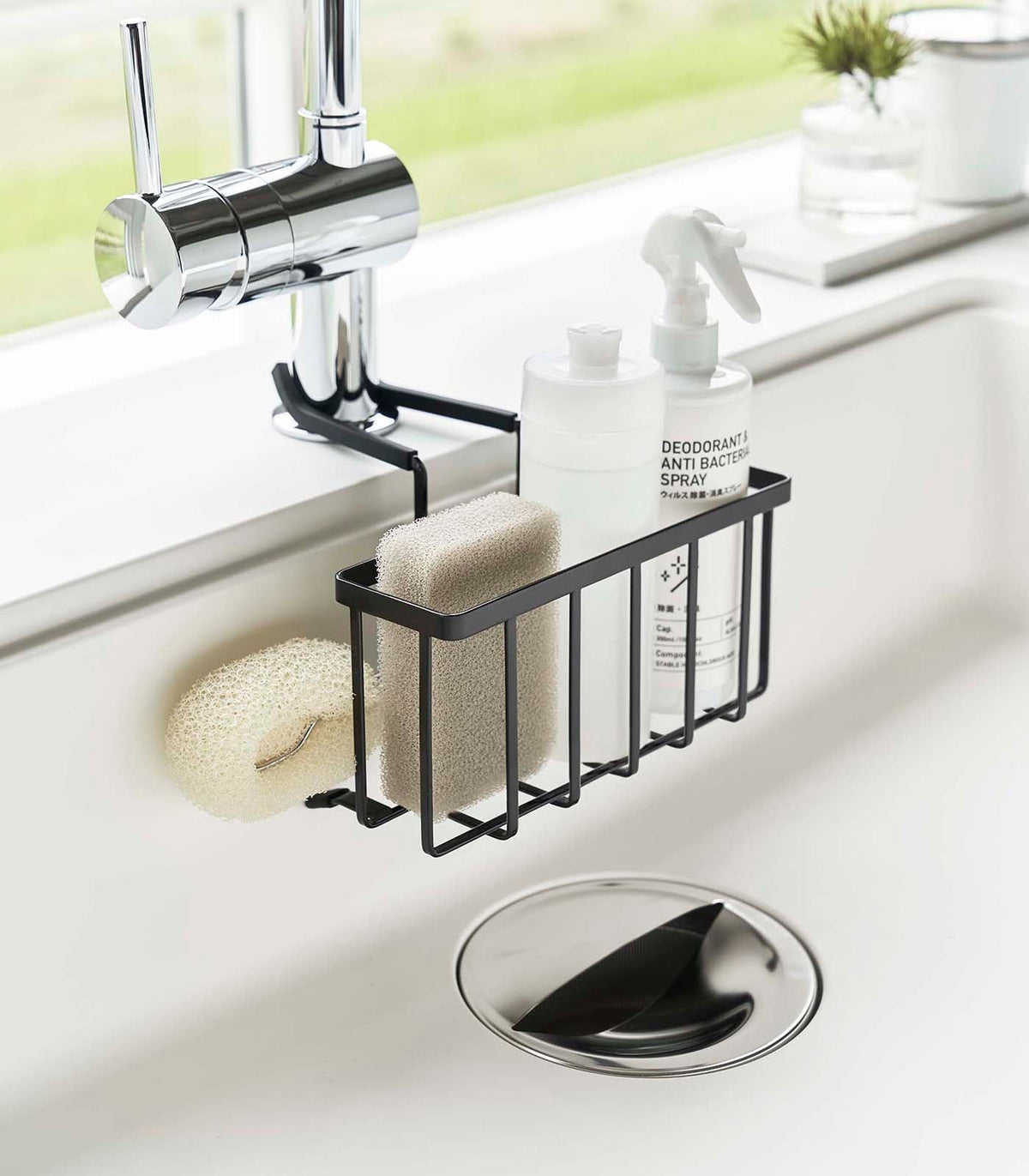 Black Yamazaki Home Faucet-Hanging Sponge Caddy - Steel inside a white sink, holding a beige sponge and white bottles, with a pale scrub brush hanging from the side.