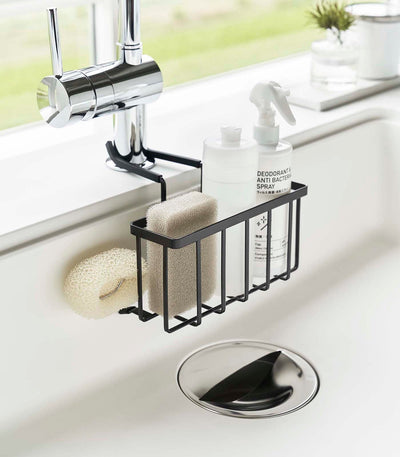 Black Yamazaki Home Faucet-Hanging Sponge Caddy - Steel inside a white sink, holding a beige sponge and white bottles, with a pale scrub brush hanging from the side.