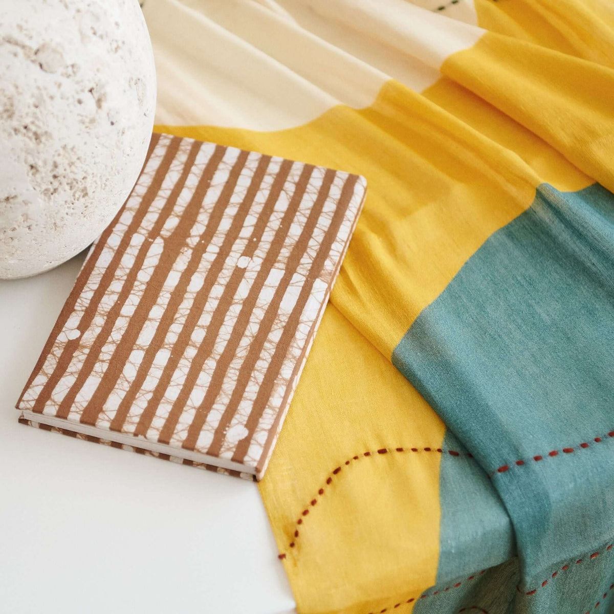 Oak Brown artisanal journal resting on layered yellow and teal textiles beside a pale ceramic object