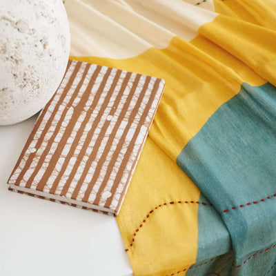 Oak Brown artisanal journal resting on layered yellow and teal textiles beside a pale ceramic object