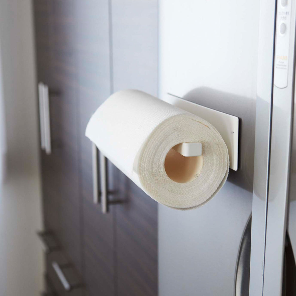 White Yamazaki Home Magnetic Paper Towel Holder - Steel mounted horizontally on a silver refrigerator, supporting a white paper towel roll with its cardboard core facing outward beside dark wood cabinets.
