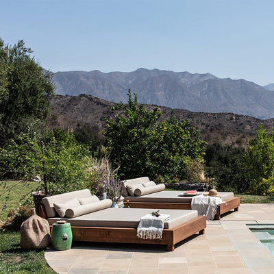 An Ojai Outdoor Daybed with cushions, placed on a tiled area next to a pool with a mountainous landscape in the background.