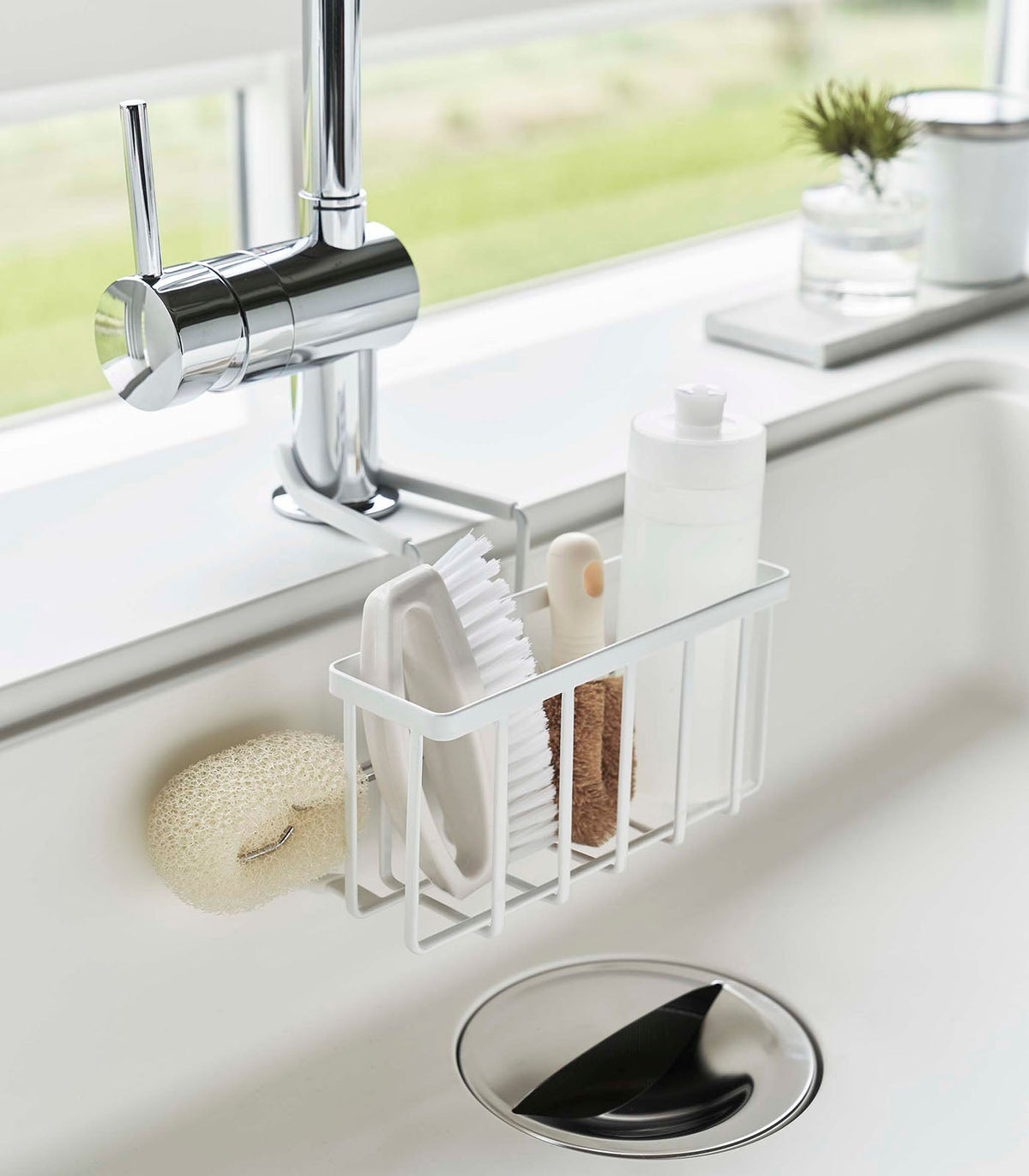 Angled view of the white Yamazaki Home Faucet-Hanging Sponge Caddy - Steel filled with bottles and cleaning tools, with a pale scrub brush hanging beside the basket above the sink drain.
