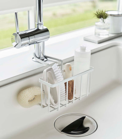 Angled view of the white Yamazaki Home Faucet-Hanging Sponge Caddy - Steel filled with bottles and cleaning tools, with a pale scrub brush hanging beside the basket above the sink drain.