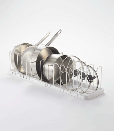 Angled view of the white Yamazaki Home Adjustable Pot Lid Organizer - Steel on a pale background, with cookware arranged from tall handled pans to smaller glass lids.