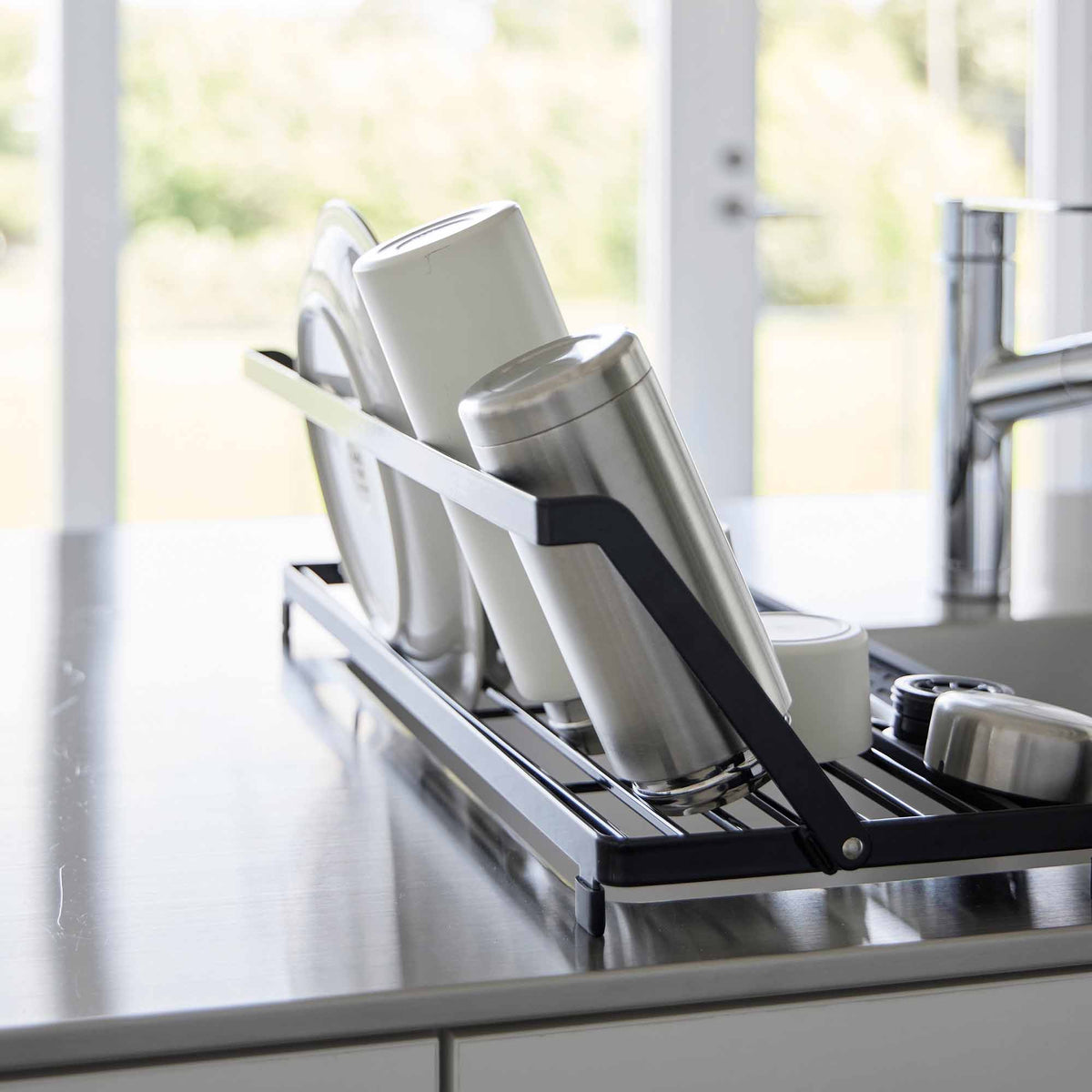 Side detail of the black Yamazaki Home Slim Over-the-Sink Dish Drying Rack - Steel, with tall white and silver-toned drinkware resting against the raised rail beside the faucet.