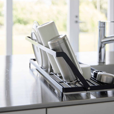 Side detail of the black Yamazaki Home Slim Over-the-Sink Dish Drying Rack - Steel, with tall white and silver-toned drinkware resting against the raised rail beside the faucet.