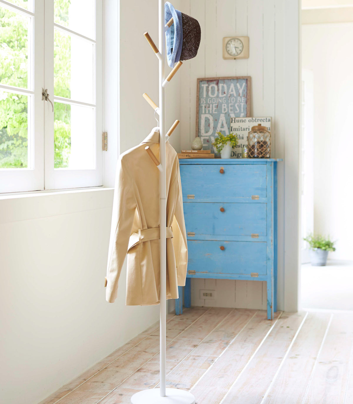 White Yamazaki Home Coat Rack (70" H) - Steel beside a sunny window, holding a pale yellow coat and blue-trimmed hat near a blue chest of drawers.