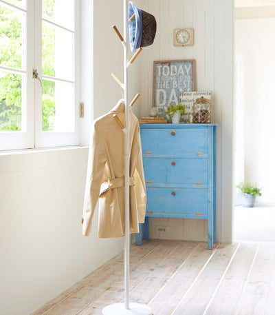 White Yamazaki Home Coat Rack (70" H) - Steel beside a sunny window, holding a pale yellow coat and blue-trimmed hat near a blue chest of drawers.