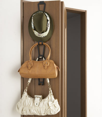 Black Yamazaki Home Cascading Storage Hooks - Steel hanging over a wooden door, holding an olive hat, tan handbag and cream handbag in a vertical arrangement.