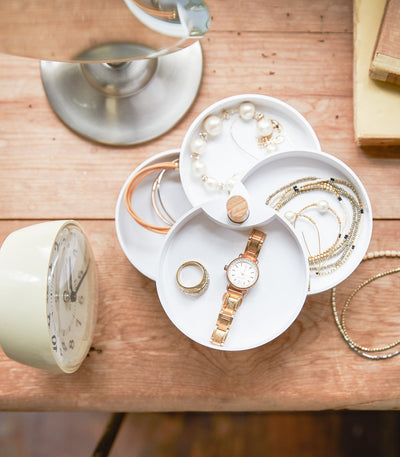 Top-down view of the Yamazaki Home Open Top Stacked Jewelry Organizer with four white trays fanned around the wooden pivot, displaying a watch, rings, bracelets and pearl-like pieces beside a small clock.