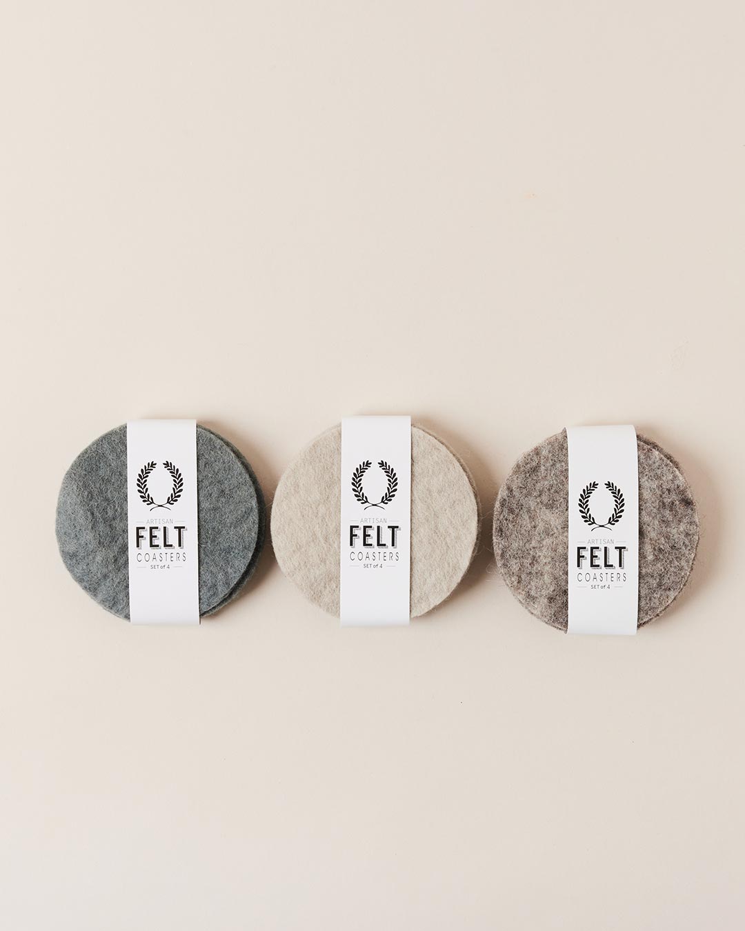 Three bundled sets of round felt coasters in charcoal gray, warm oatmeal, and mottled taupe, each wrapped with a white paper band labeled “Felt Coasters.”