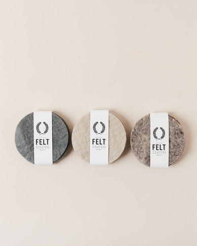 Three bundled sets of round felt coasters in charcoal gray, warm oatmeal, and mottled taupe, each wrapped with a white paper band labeled “Felt Coasters.”
