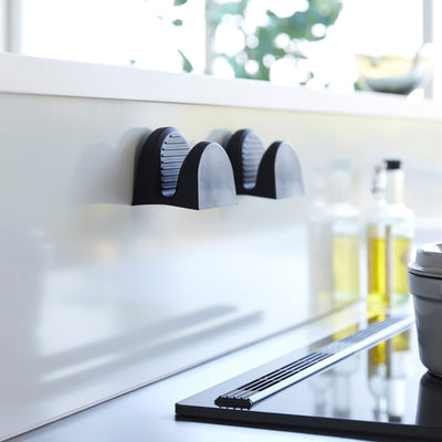 Two black Yamazaki Home Magnetic Pot Holders - Set of 2 - Silicone attached side by side to a pale kitchen backsplash, with their ridged interiors visible.
