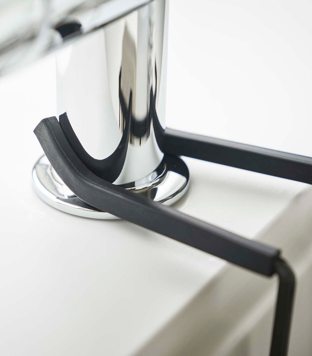 Close-up of the black Yamazaki Home Faucet-Hanging Sponge Caddy - Steel arms resting around the chrome faucet base and extending over the pale sink rim.