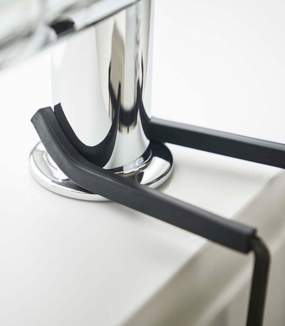 Close-up of the black Yamazaki Home Faucet-Hanging Sponge Caddy - Steel arms resting around the chrome faucet base and extending over the pale sink rim.