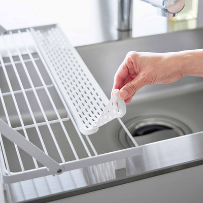 A hand lifts the perforated utensil tray from the white Yamazaki Home Slim Over-the-Sink Dish Drying Rack - Steel, revealing the open bars beneath it over the sink.