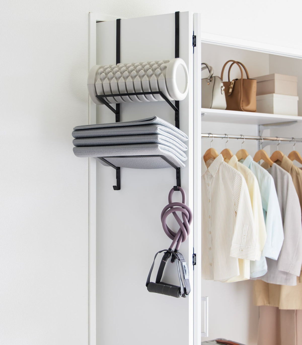 Black Yamazaki Home Over-the-Door Fitness Equipment Hanger - Steel storing a grey foam roller, folded grey mat and purple resistance bands on a closet door beside hanging clothes.