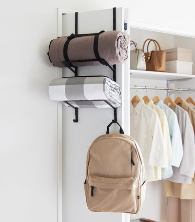 Black Yamazaki Home Over-the-Door Fitness Equipment Hanger - Steel used on a wardrobe door to hold a rolled brown blanket, folded grey-and-white textile and tan backpack.
