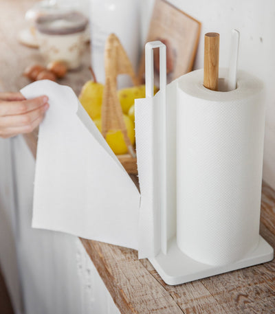 Wider kitchen view of a person drawing a sheet from the Yamazaki Home Paper Towel Holder - Steel + Wood, with its white rectangular base, tall side rail and wooden post visible beside lemons and cutting boards.
