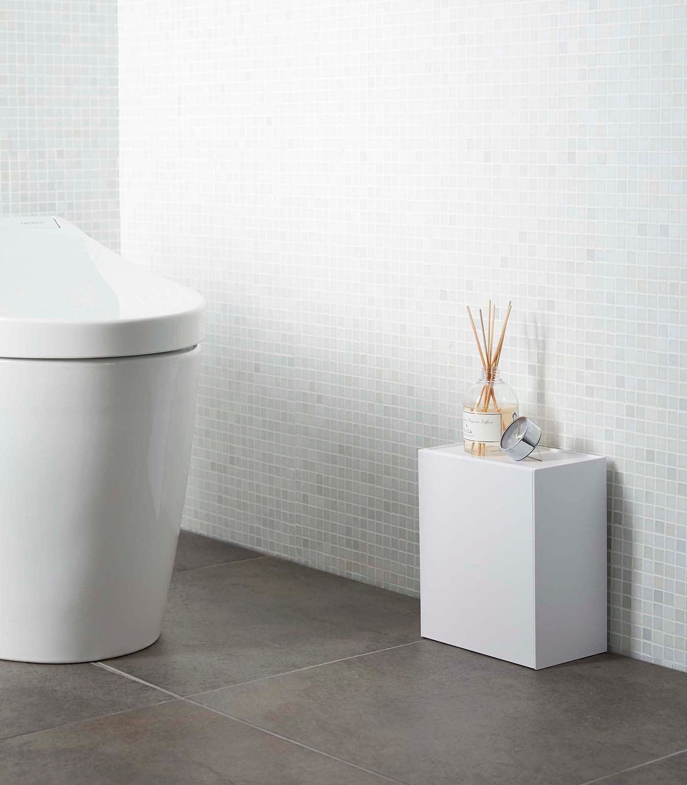 Modern bathroom with a toilet and small white cabinet against a tiled wall. Wall-Mounted Storage or Trash Bin shown on floor.