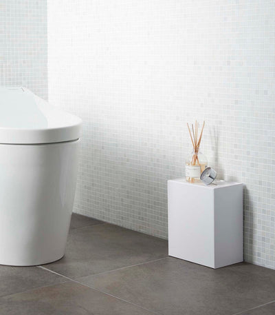 Modern bathroom with a toilet and small white cabinet against a tiled wall. Wall-Mounted Storage or Trash Bin shown on floor.
