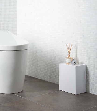 Modern bathroom with a toilet and small white cabinet against a tiled wall. Wall-Mounted Storage or Trash Bin shown on floor.