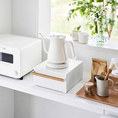 White Yamazaki Home Countertop Rack with Drawer - Steel + Wood beneath a kettle beside a compact oven, with its drawer closed and light wood handle facing forward in a bright kitchen.