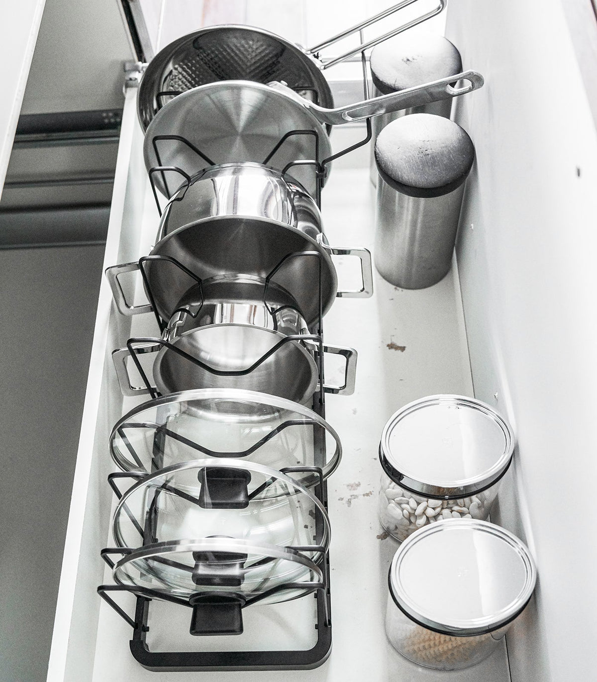 A hand lifts frying-pan handles from the black Yamazaki Home Adjustable Pot Lid Organizer - Steel while the remaining pans and lids stand between its wire dividers.