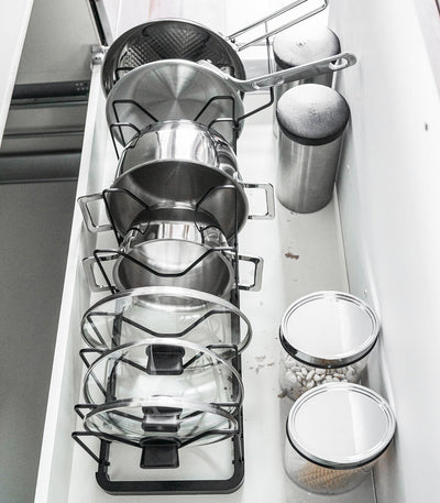 A hand lifts frying-pan handles from the black Yamazaki Home Adjustable Pot Lid Organizer - Steel while the remaining pans and lids stand between its wire dividers.