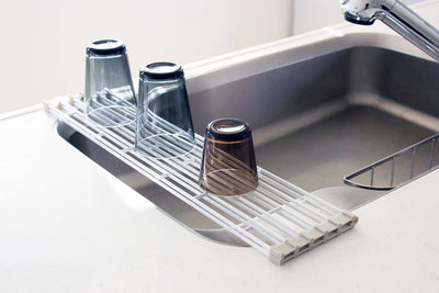 White Yamazaki Home Over-the-Sink Dish Drainer - Steel partially covering a stainless-steel sink, with three inverted blue and brown drinking glasses resting on its parallel bars.