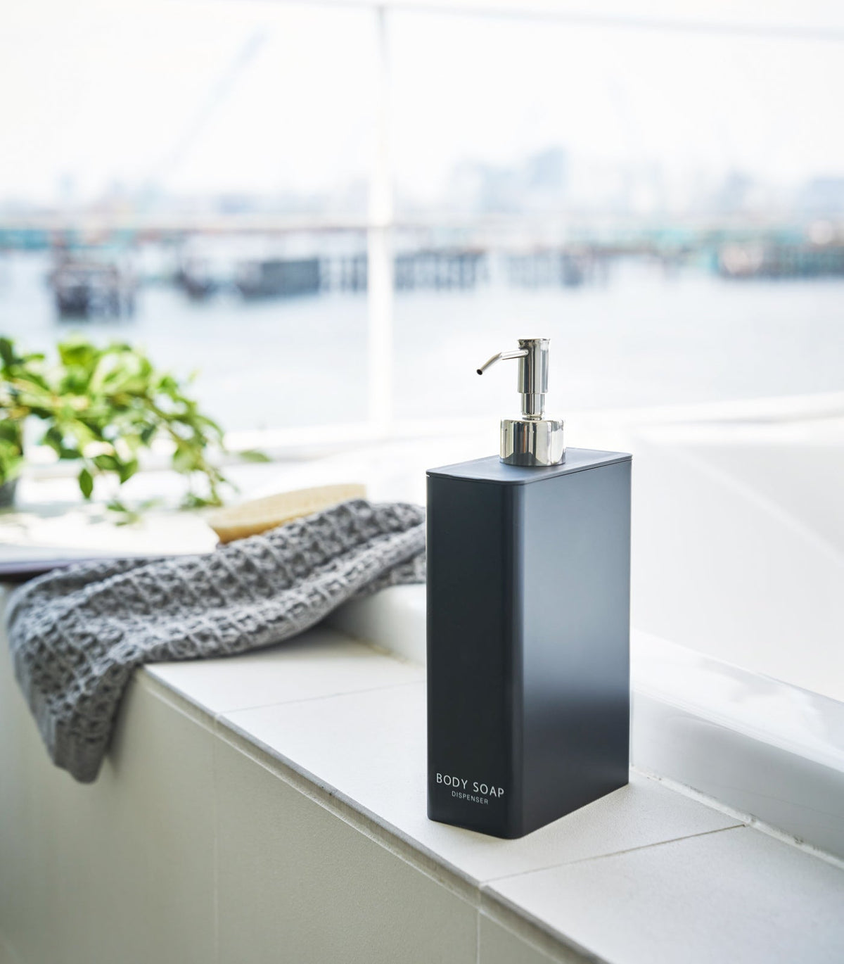 Black Yamazaki Home Rectangle Shower Dispenser - Three Styles labelled BODY SOAP on a white bathtub ledge, with a silver pump, folded grey towel and leafy plant nearby.